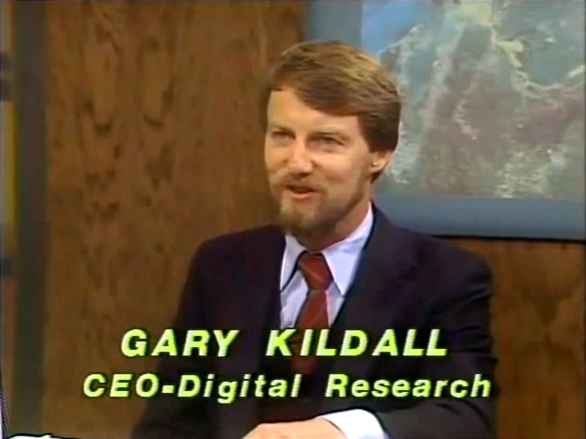 Gary Kildall on the other side of the desk, looking dapper in a dark blue jacket, light blue dress shirt, and burgundy tie with a wide knot.