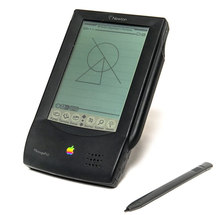 An Apple Newton MessagePad 100 sits at a jaunty angle on a white table, flat, stubby stylus beside it. On screen, a circle, line, and triangle are drawn in the Notes app. I just realized this unit is in German.