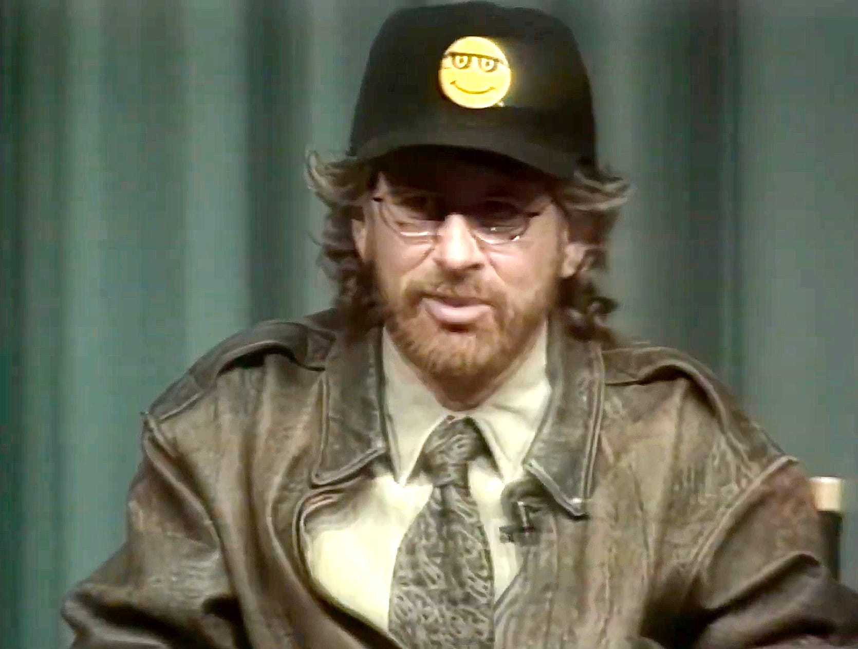 Screenshot from Triumph of the Nerds. Steven Spielberg, in a heavy, brown leather jacket, yellow shirt, wide brown tie wears a black hat with Microsoft Bob's yellow smiling face on it.