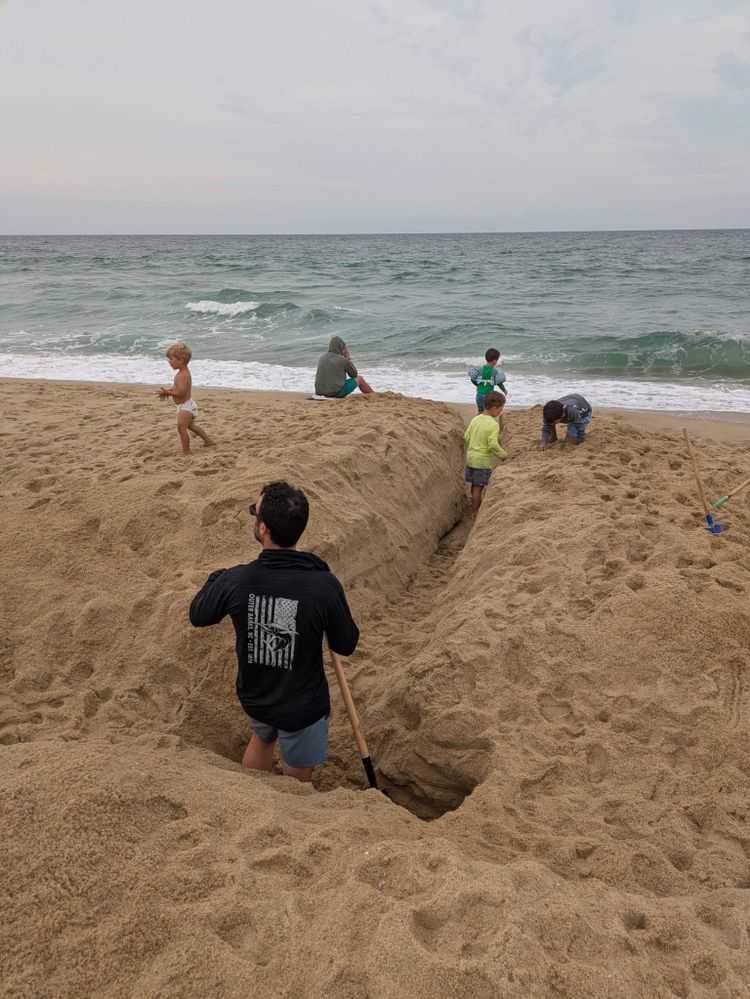 Steve waist-deep in a sand trench aimed at the ocean, kids digging alongside
