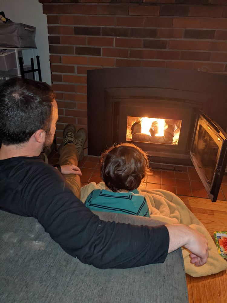 A father's shoulder and his son under a blanket watching a fire, boots by the hearth