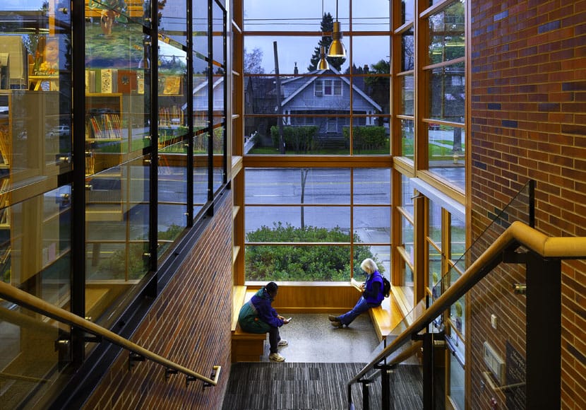 A view from the interior of Montlake Library looking at a reading nooks with two readers at it and a single family home across the street.