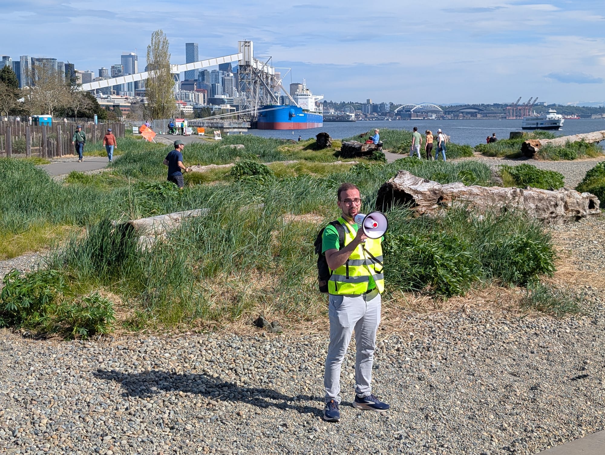 The Seattle skyline and Elliott Bay is in the background at Carl speaks with his yellow safety vest and megaphone. 