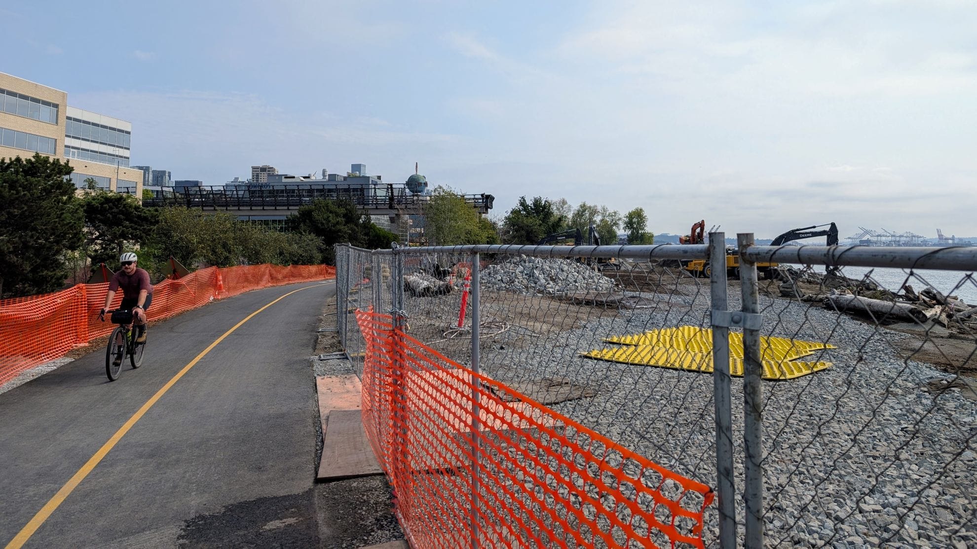 A person bike in the Elliott Bay Trail with orange fencing and chainlink separating the construction zone where a backhoe is at work. 