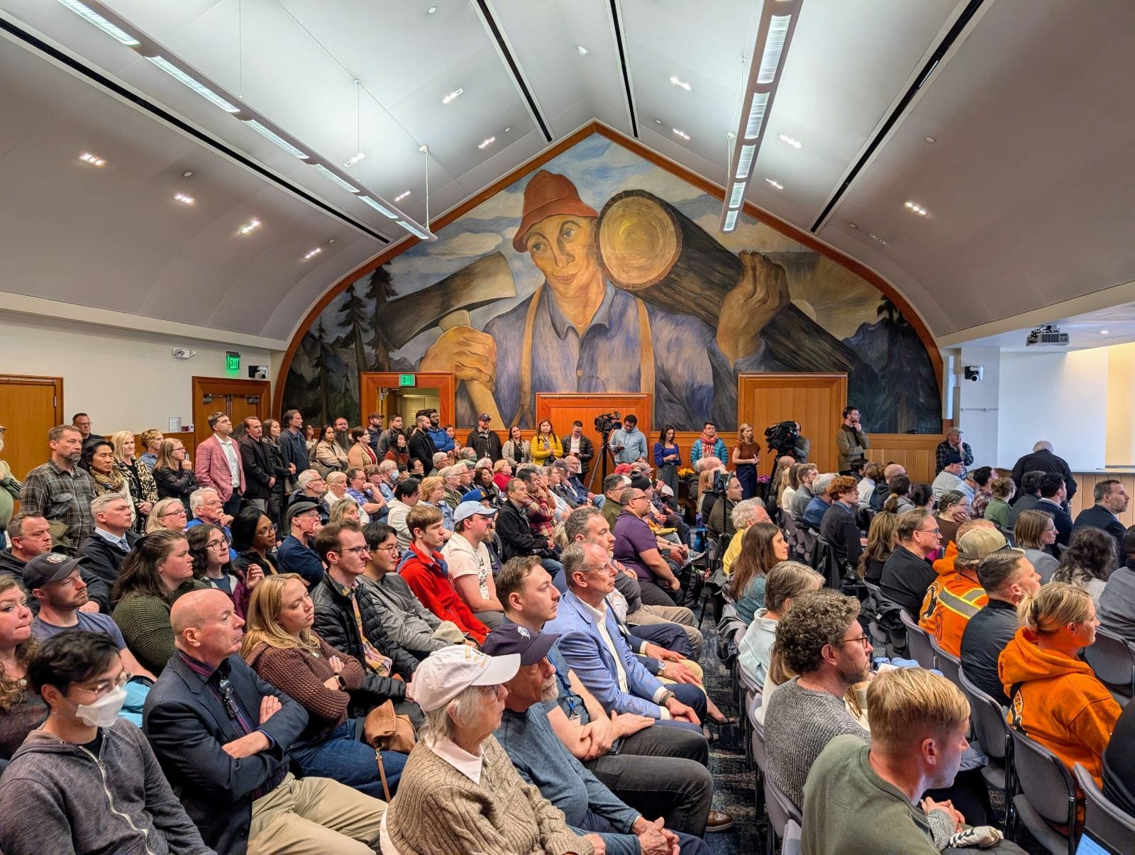 A giant lumberjack mural grips an axe and a big log in a mural on the north wall. More than 100 people crowd the room to watch the panel discussion.