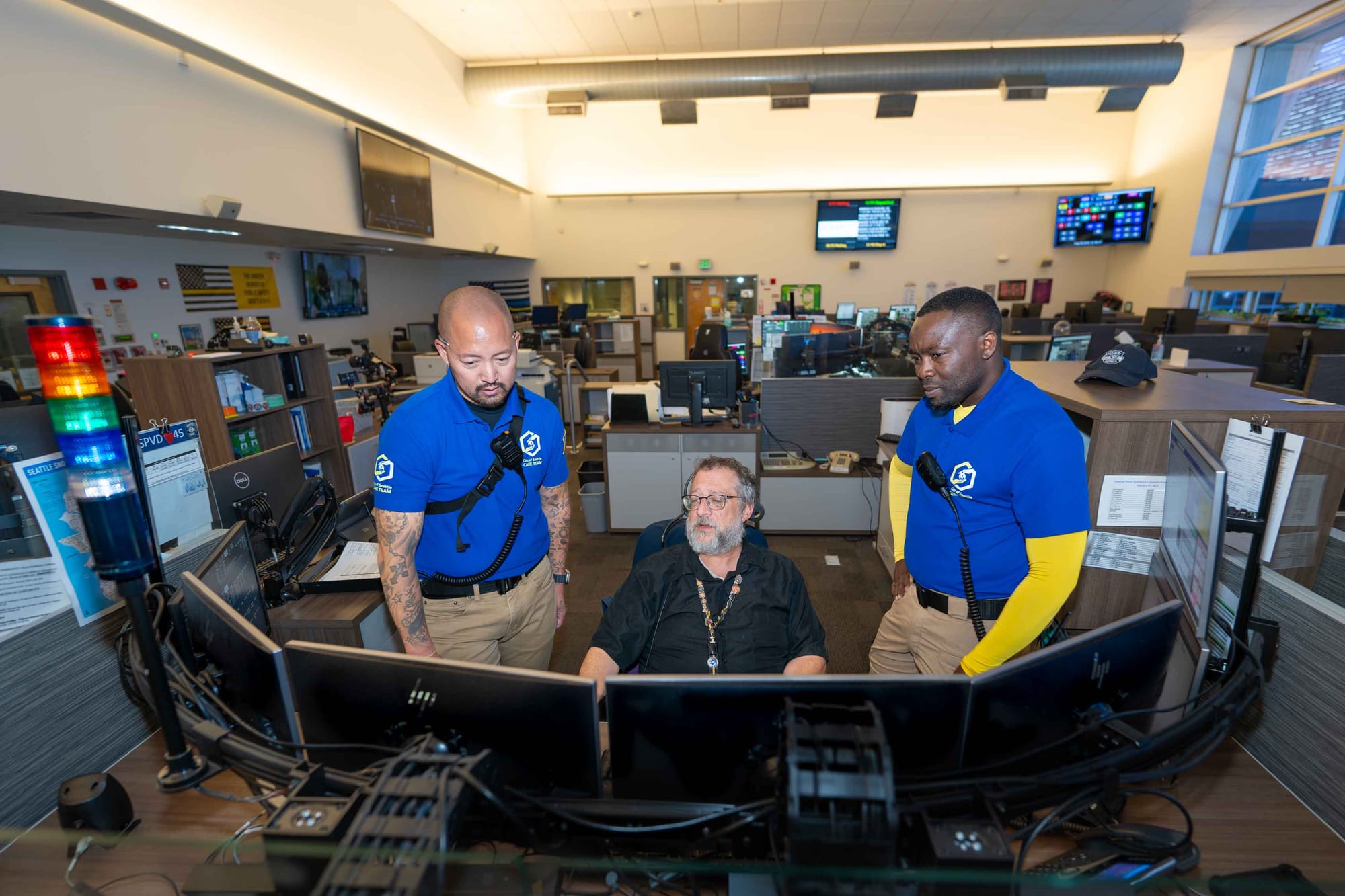 Two CARE responders in blue uniforms stand next to a dispatcher sitting around a number of monitors in an office.