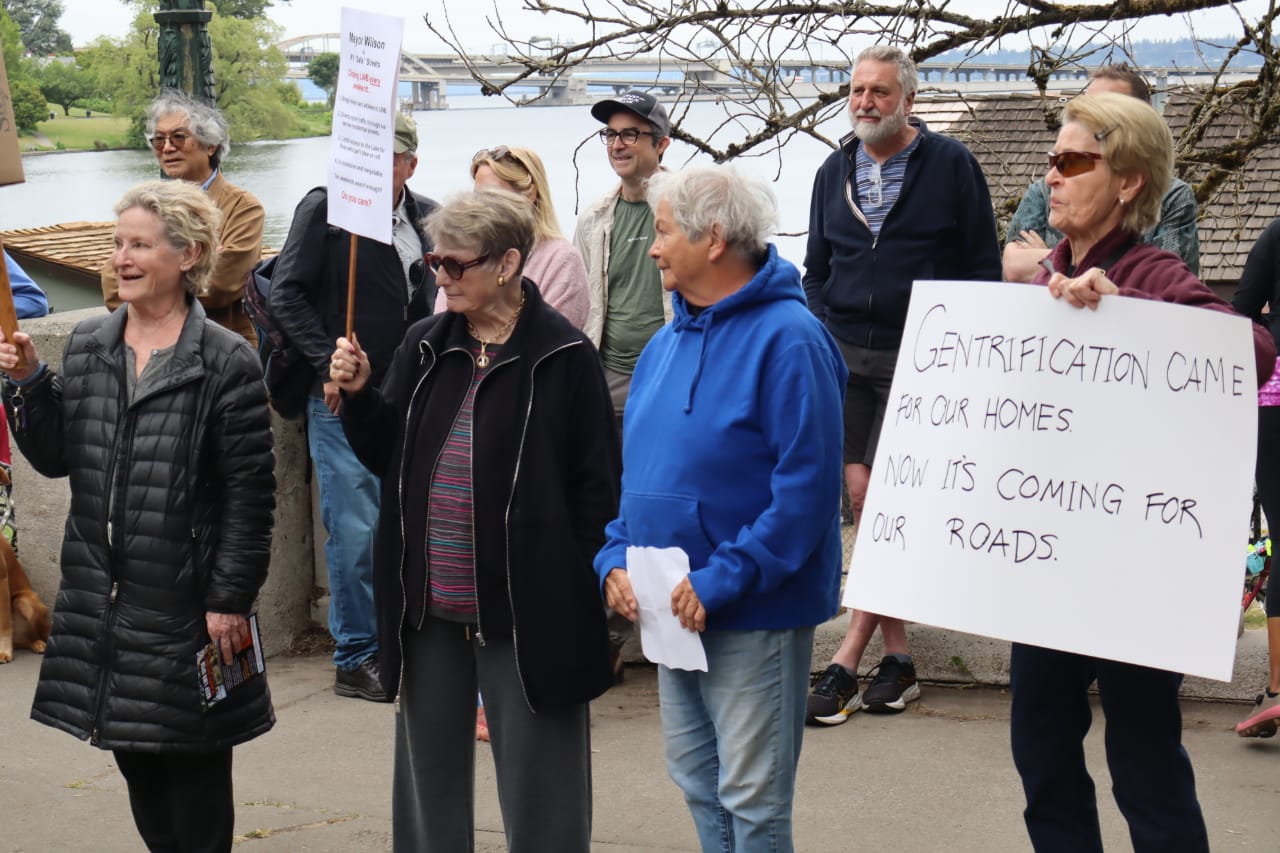A group of mostly senior citizens holds protest signs. One reads "Gentrification came for our homes. Now it's coming for our roads."