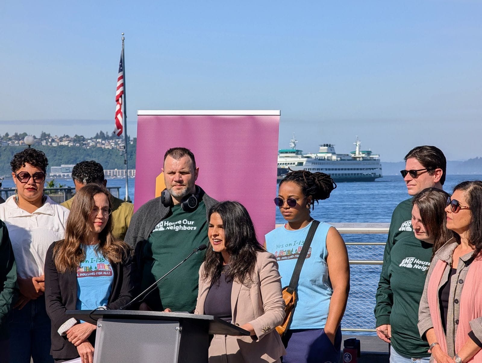 Rinck stands at the lectern with a Washington State ferry pulling into the bay in the background.