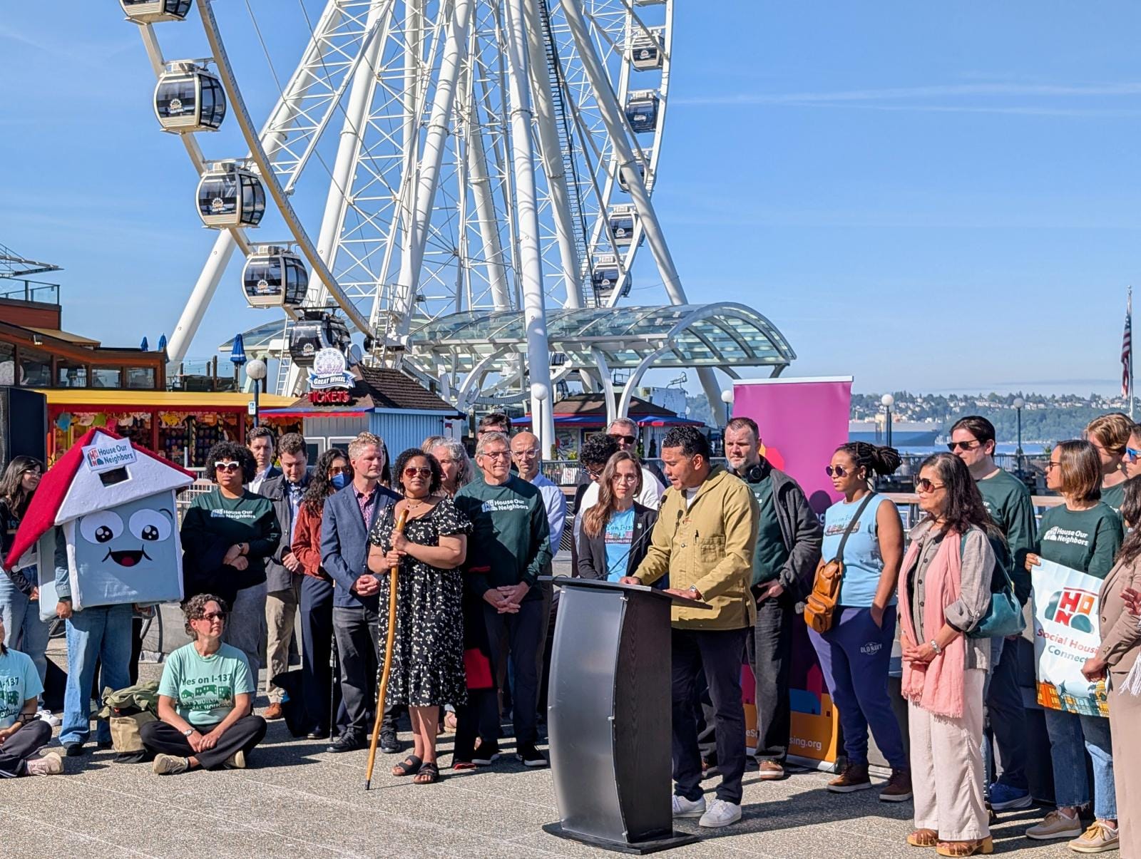 Three dozen supporters stand behind speaks at Pier 58 with the Ferris wheel and Elliott Bay in the background.