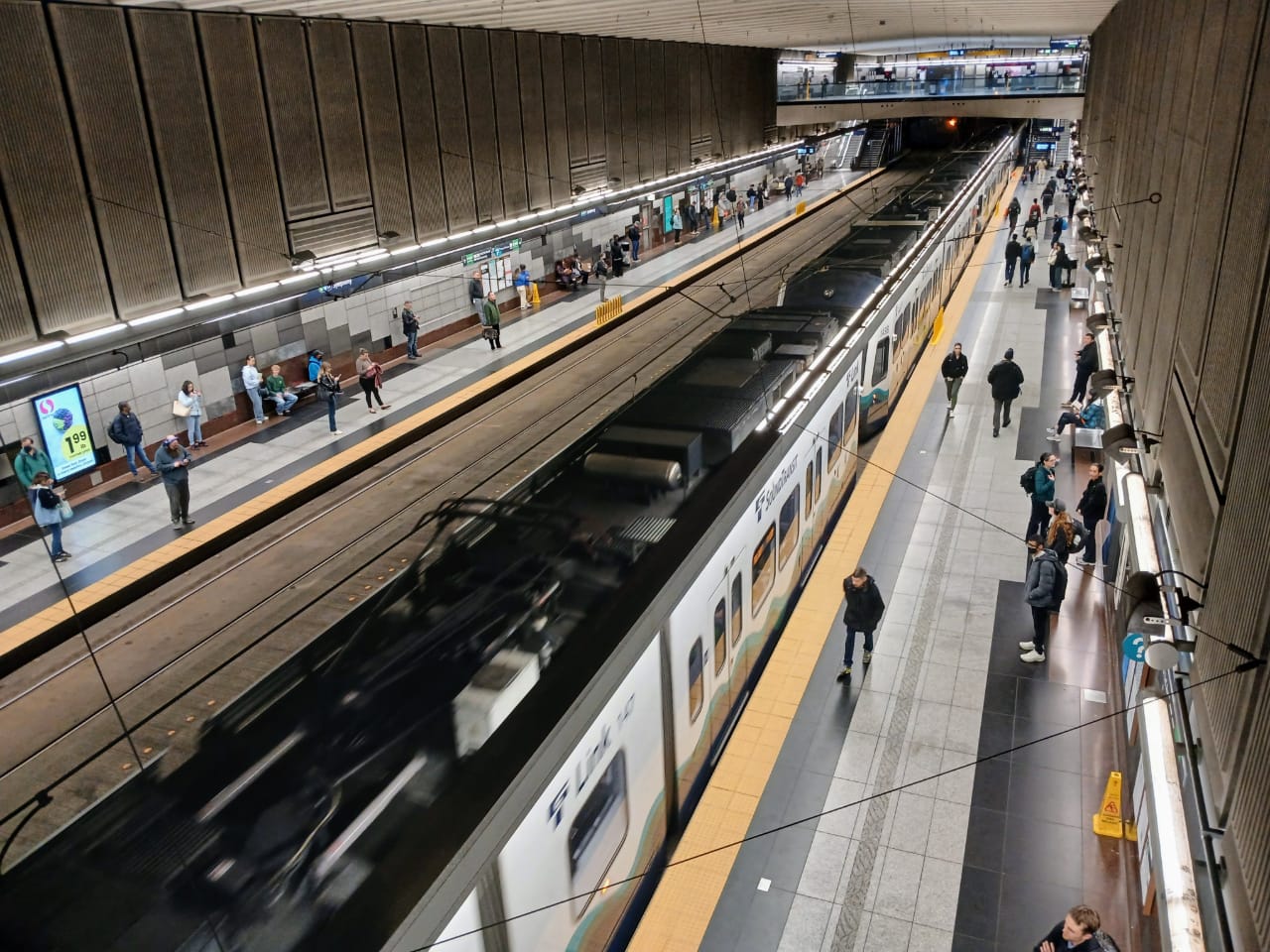 A view looking down on the station platform from the mezzanine with upwards of a 100 passengers spread around the station and a train pulling up.