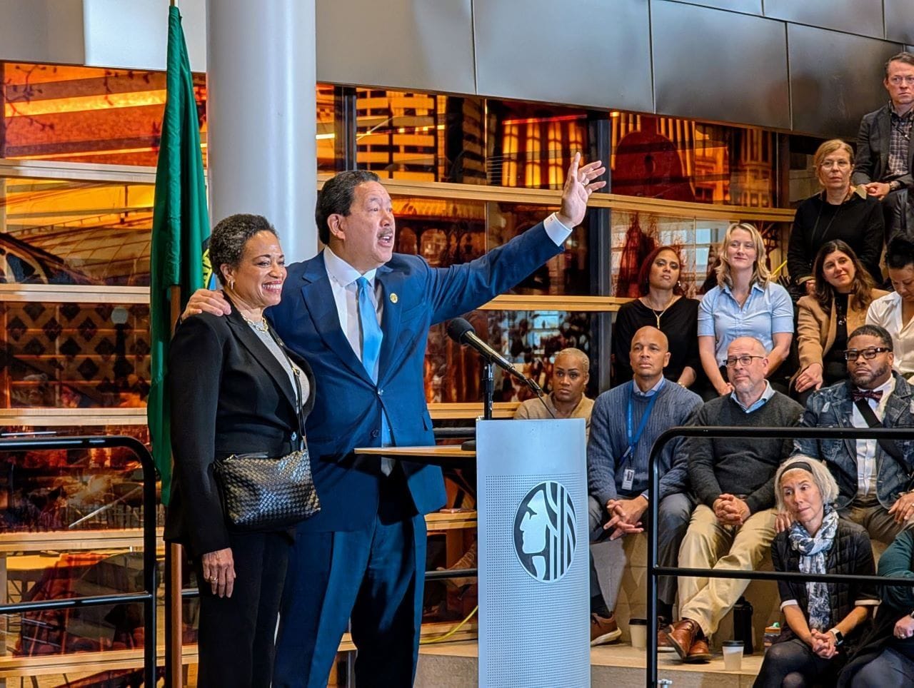 Wearing a suit, Bruce Harrell stand at a lectern and waves to the crowd with one hand which gripping his wife's shoulder with another.