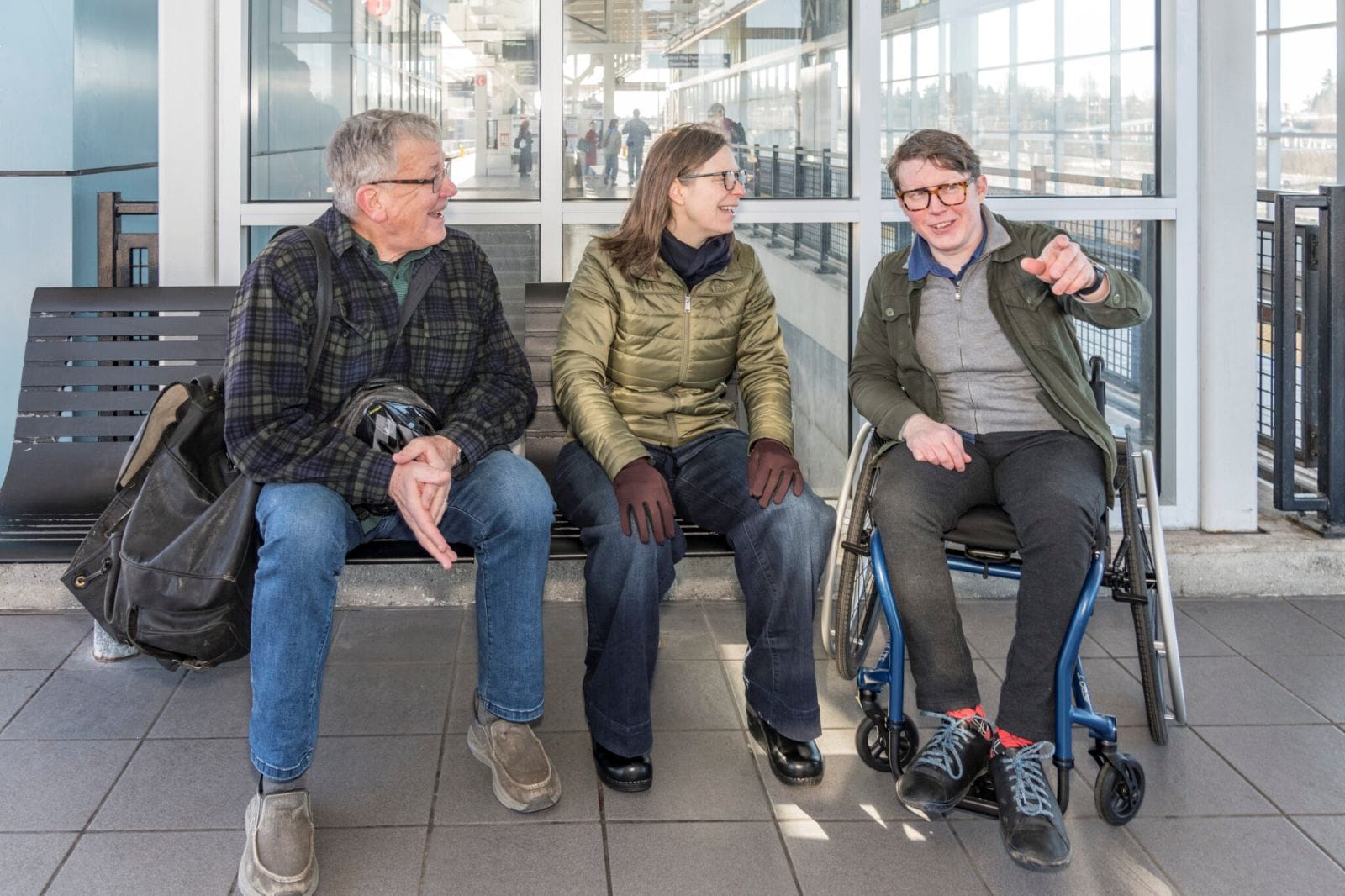 Silas is in a wheelchair and talking two people sitting on a bench at an elevated light rail station.