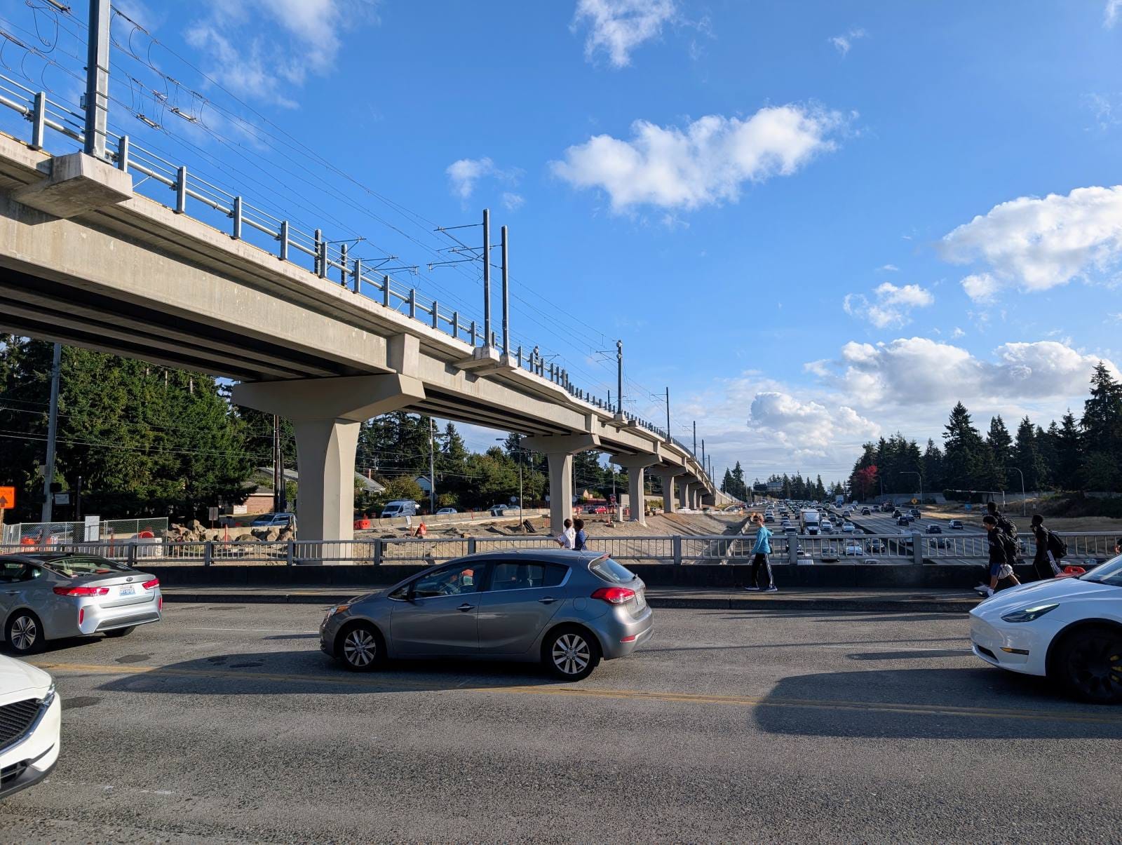 The view south of NE A car-choked 130th Street with the elevated guideway and freeway trench. A few kids walk on the overpass sidewalk