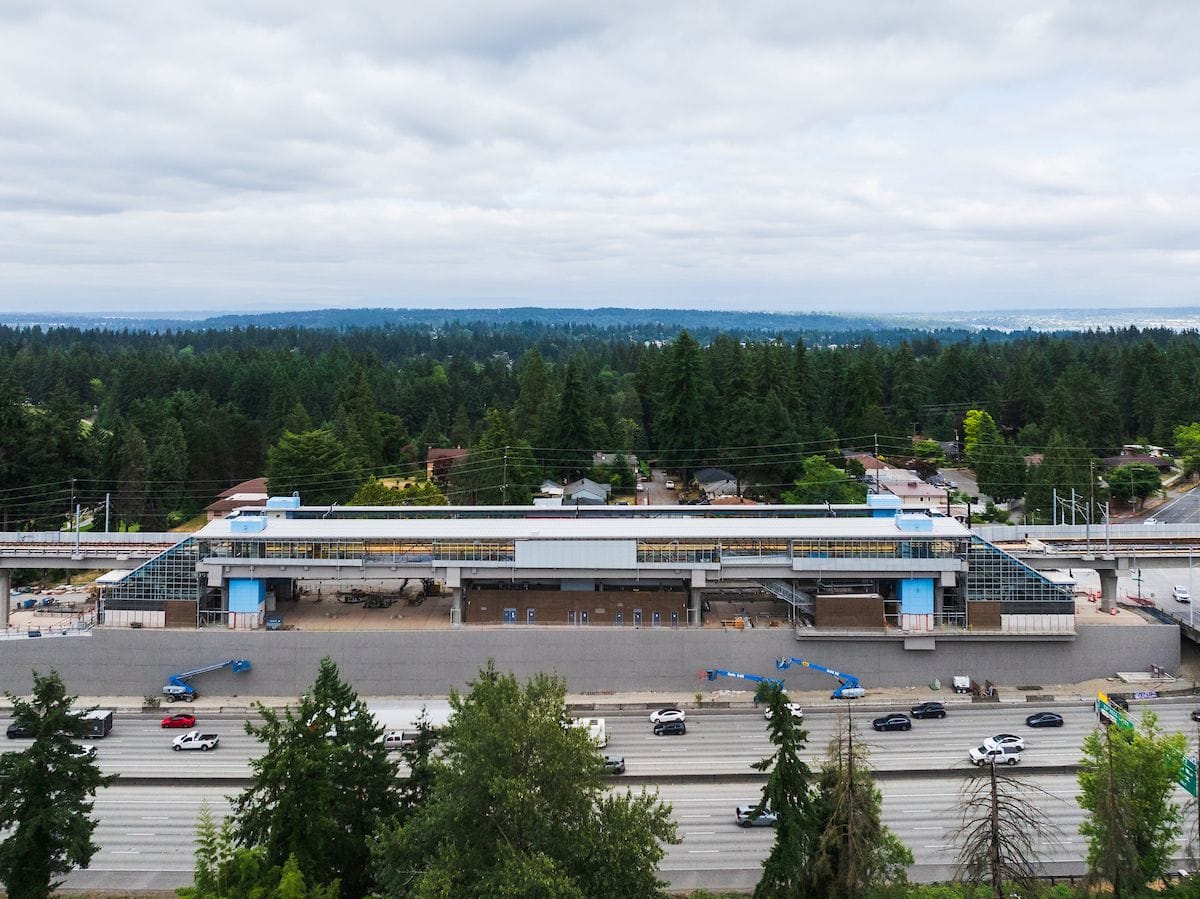 An aerial drone shot shows I-5 in the foreground, the elevated station in the midground, and tons of tree tops, but little housing visible in the distance.