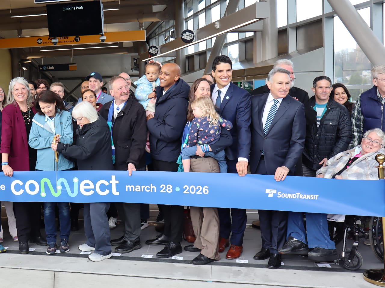 Constantine wears a suit and stands next to former Sound Transit CEO Joni Earl. To the left Katie Wilson and Girmay Zahilay stand along the ribbon, both holding their toddler-age daughters. US Senators Patty Murray and Maria Cantwell jointly clutch a big gold pair of ceremonial scissors for the ribbon.
