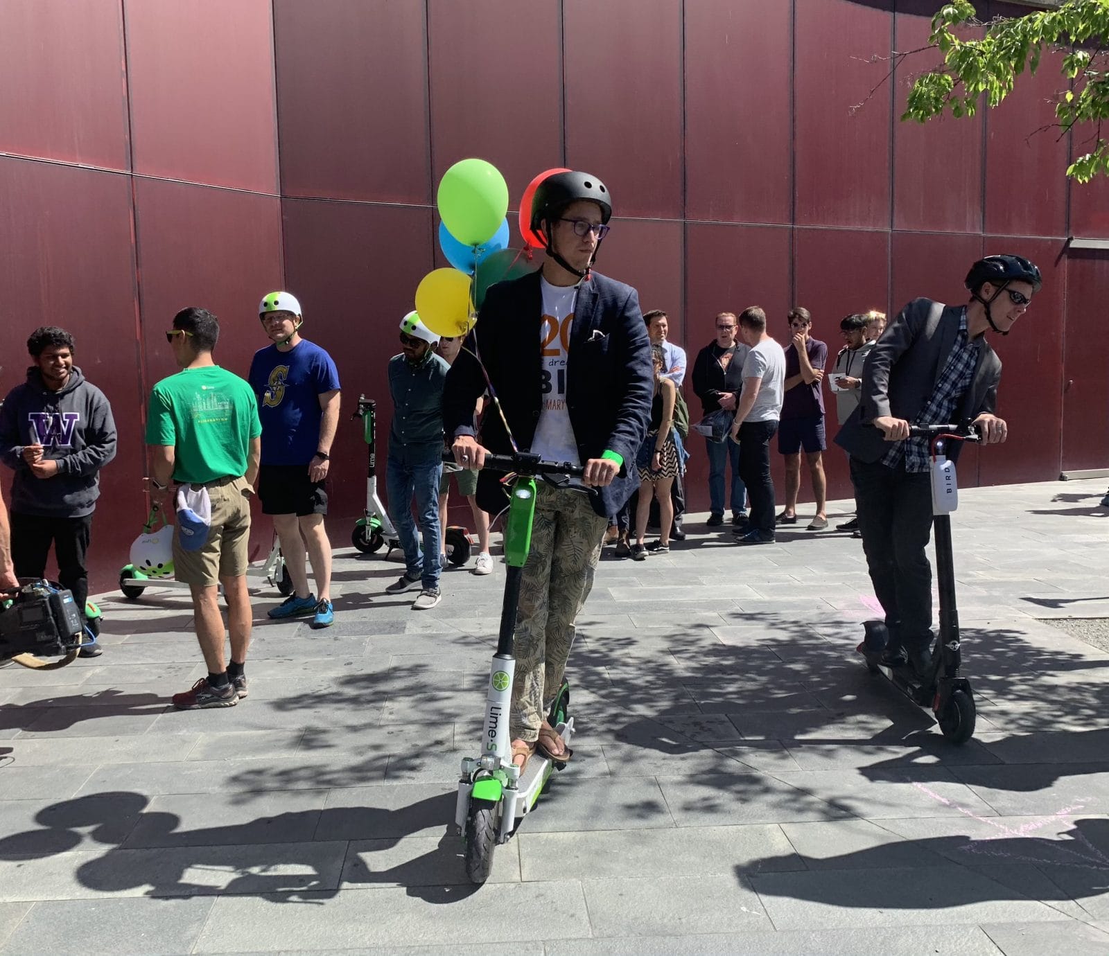 A dozen folks stand around as two people test ride e-scooters. Balloons adorn the city hall plaza and its burgundy paneled wall.