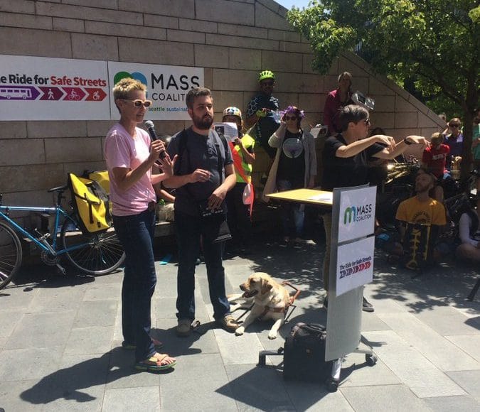 Anna Zivarts hold the microphone at a podium next to a bearded man with a guide dog with a crowd of people in bicycle helmets behind. Signs read "MASS Coalition" and "Ride for Safe Streets"