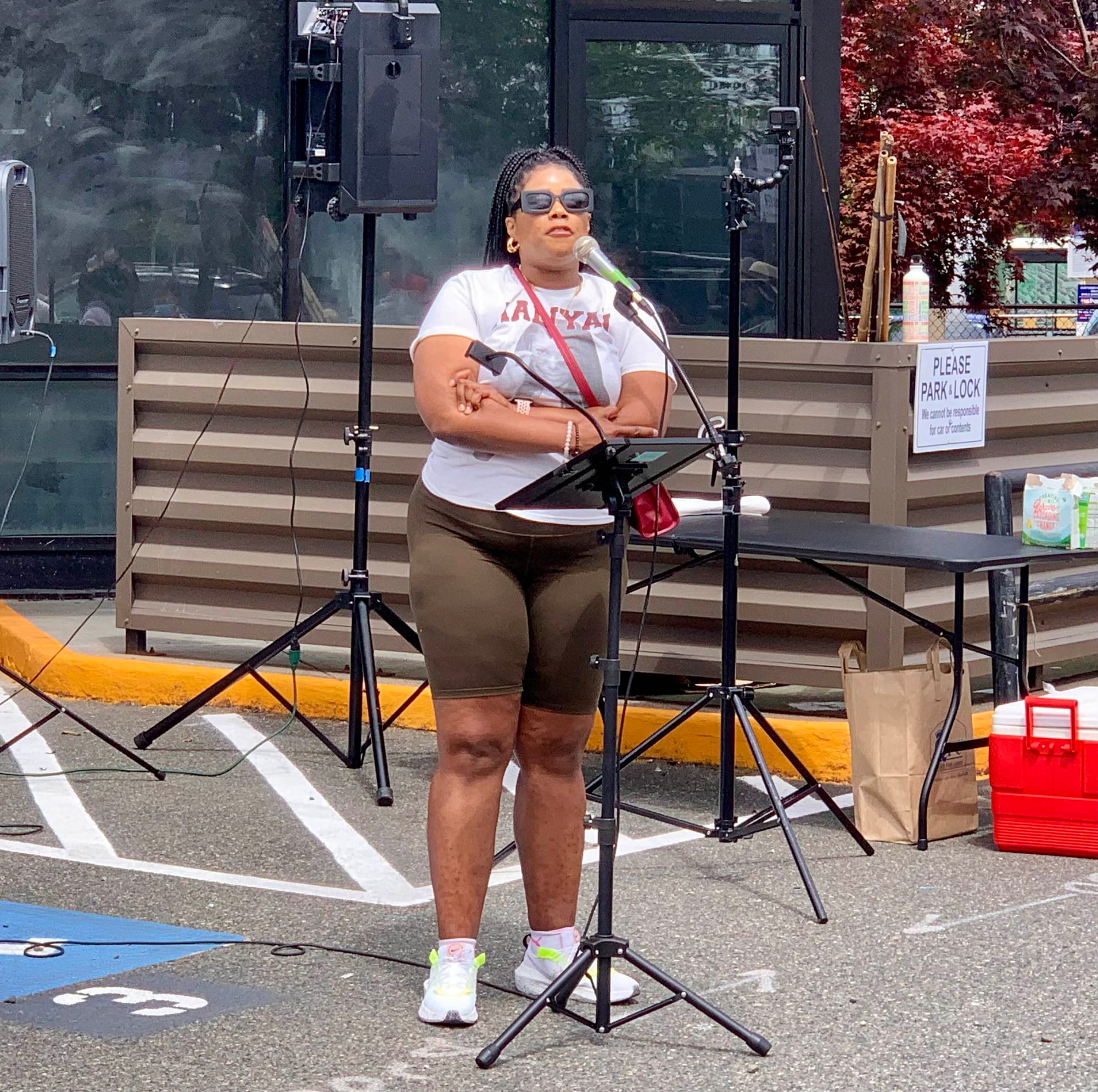 A Black woman wearing sunglasses and a red purse speaks at a podium.