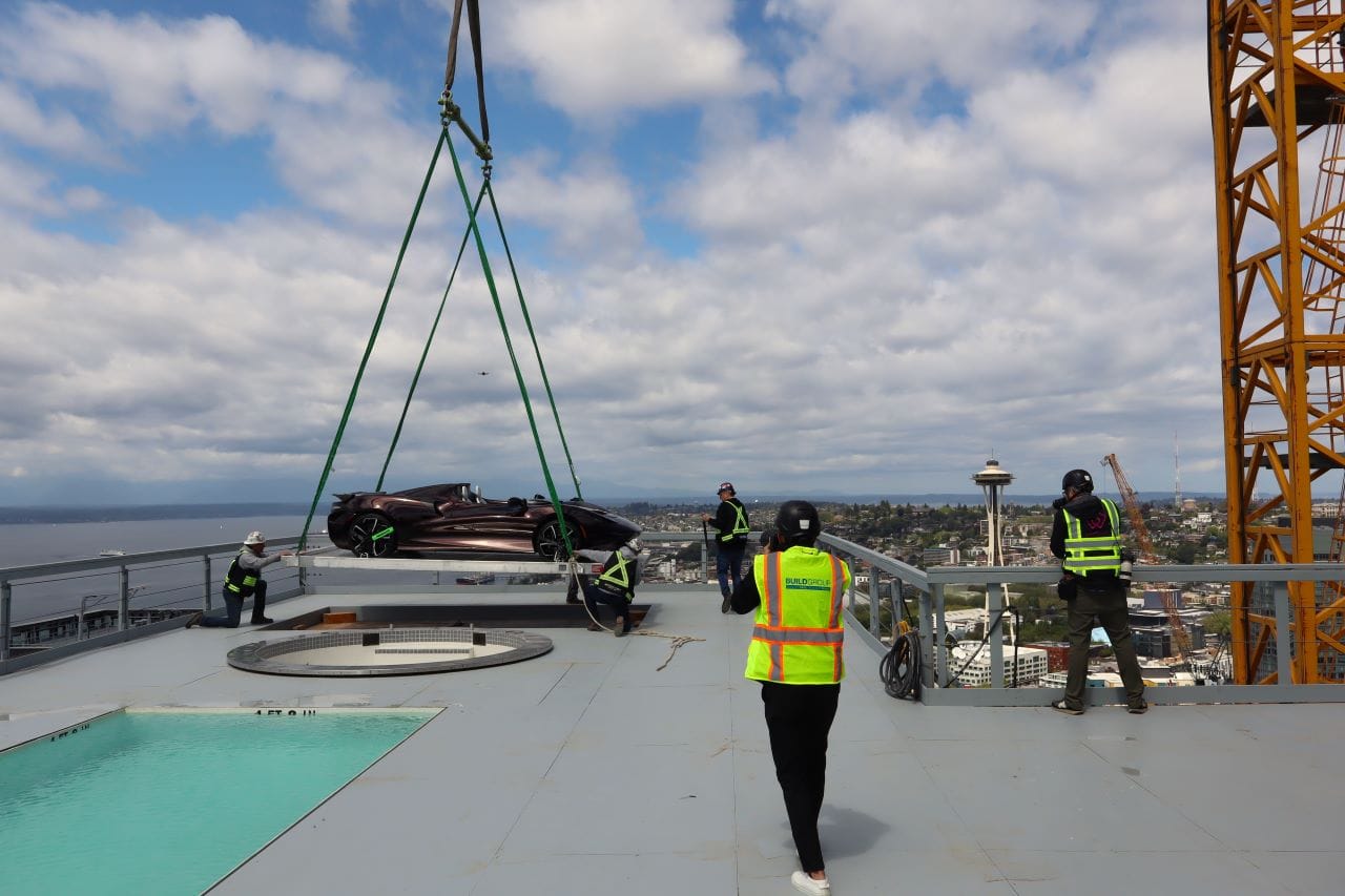 A crane lowers the sport car at the edge of the hottub with the Space Needle and uptown view in the background.