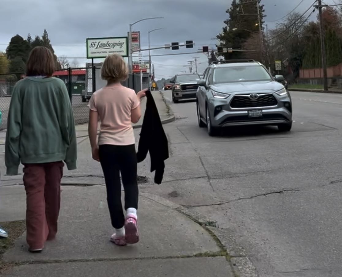 Two girls walk down 6th narrow sidewalk as cars zip by. One draps a jacket over her shoulder.