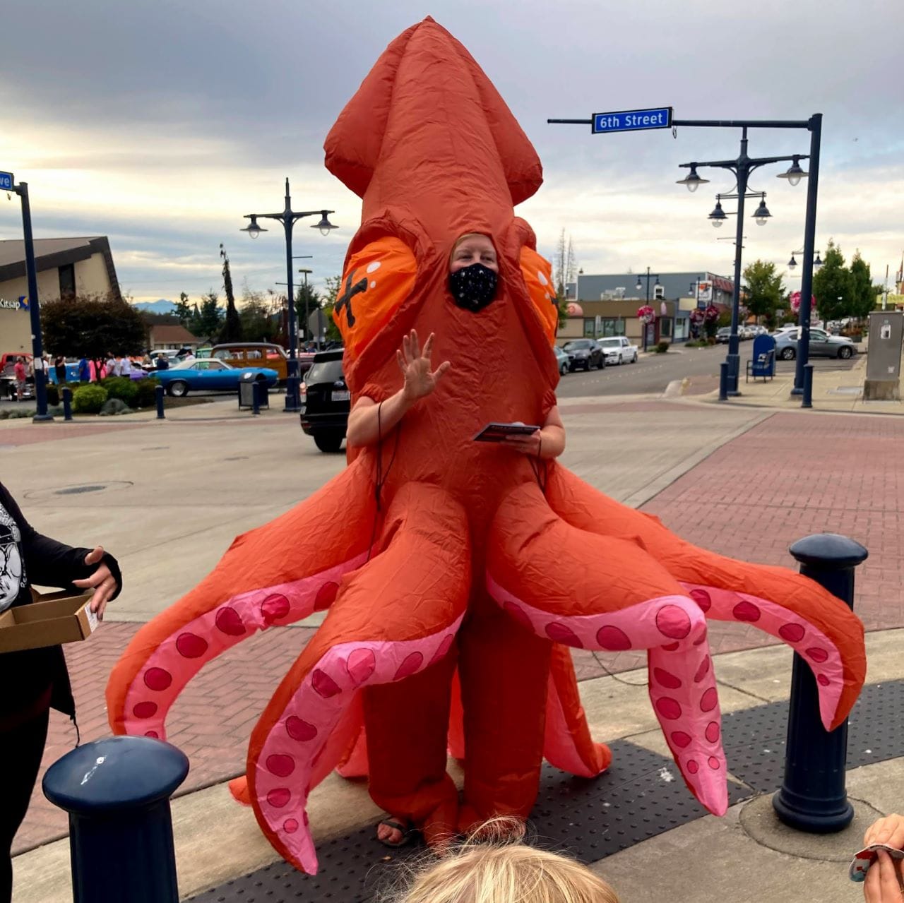 A woman in an octopus costume stands nexts to bollards at a 6th avenue intersection and gestures and reads a story to kids. Cars zip through the intersection.