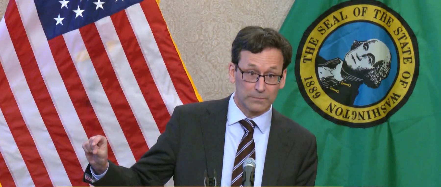 Ferguson wears a suit and glasses and gestures his figures with a little bit sign. He stands in front of the USA and Washington state flags.