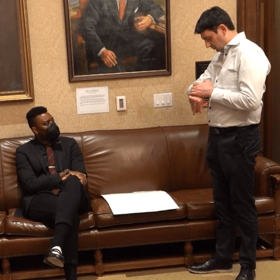 Shane Esquibel answers a call coming through on his smartwatch, as Scott sits and looks in from the Governor's waiting room, adorned with official portraits of governors long past.