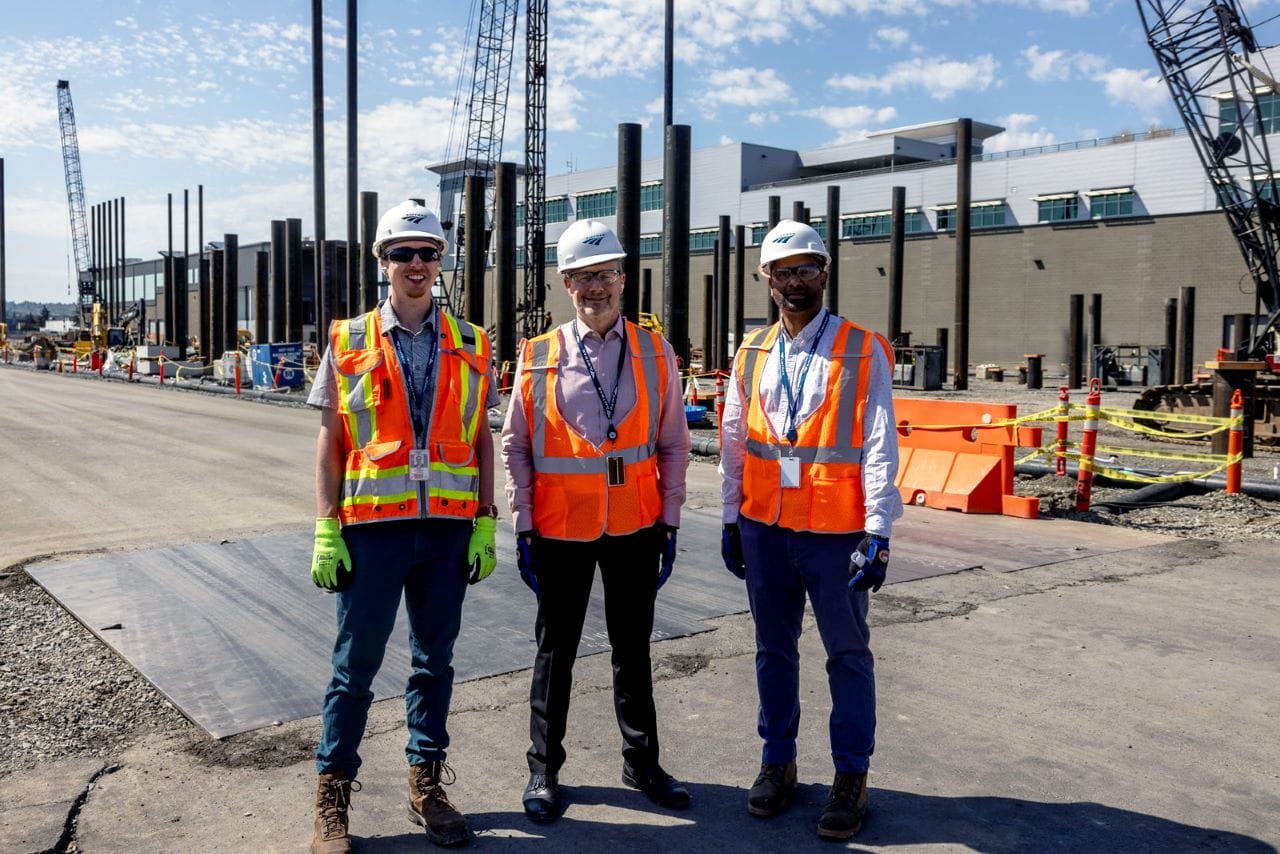 Three men pose in hard hats and orange vests with framing girders behind them for the maintenance facility.