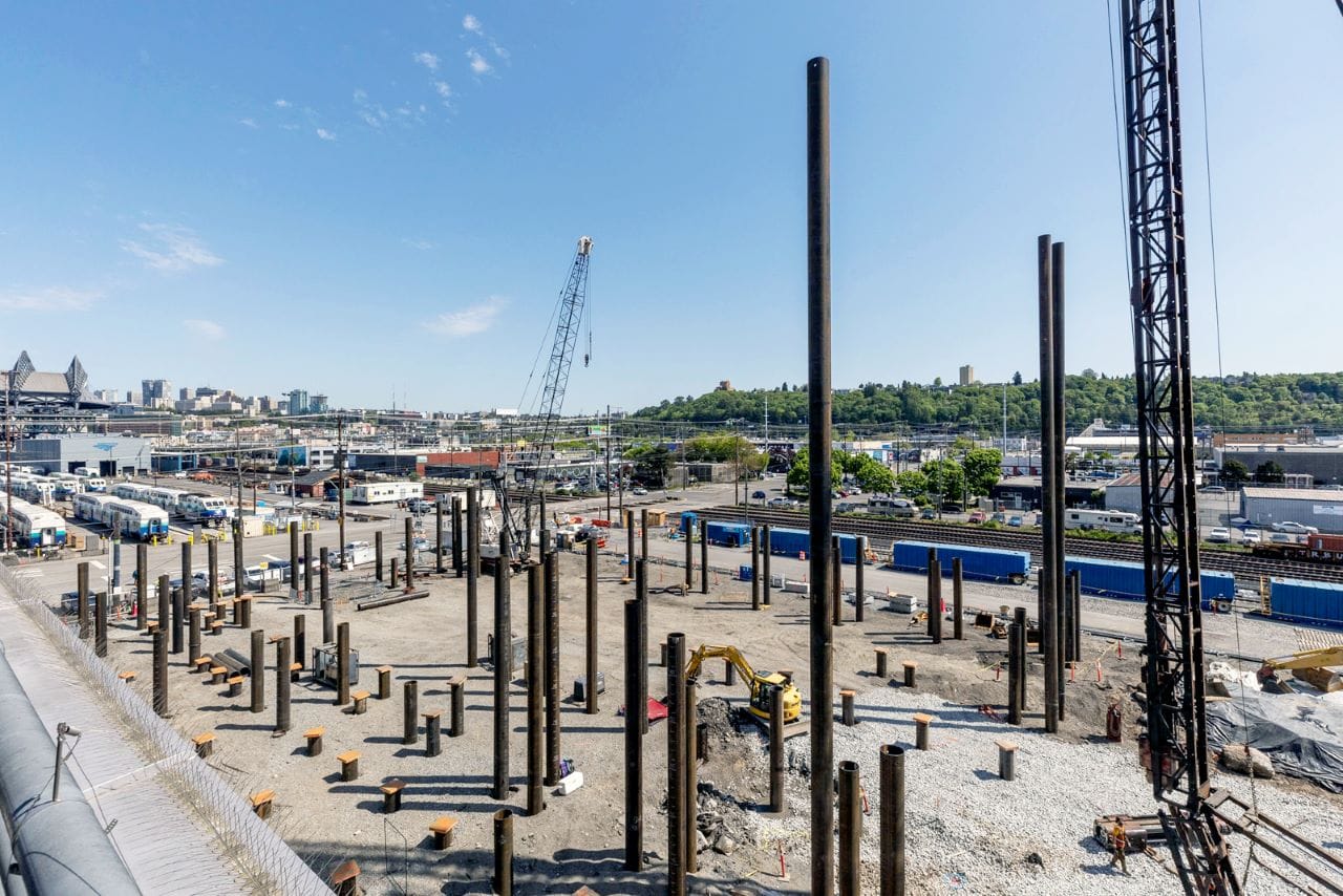 Cranes and backhoes begin work on the new facility with Sounder trains in the yard beyond and the Mariners stadium in the distance.