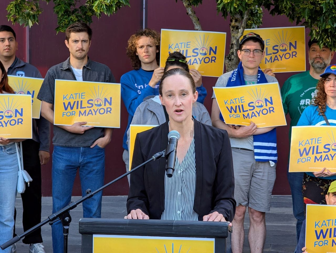 Wilson stands at a lectern speaking and a dozen supporters stand behind her holding yellow Wilson for Mayor signs on the City Hall Plaza.