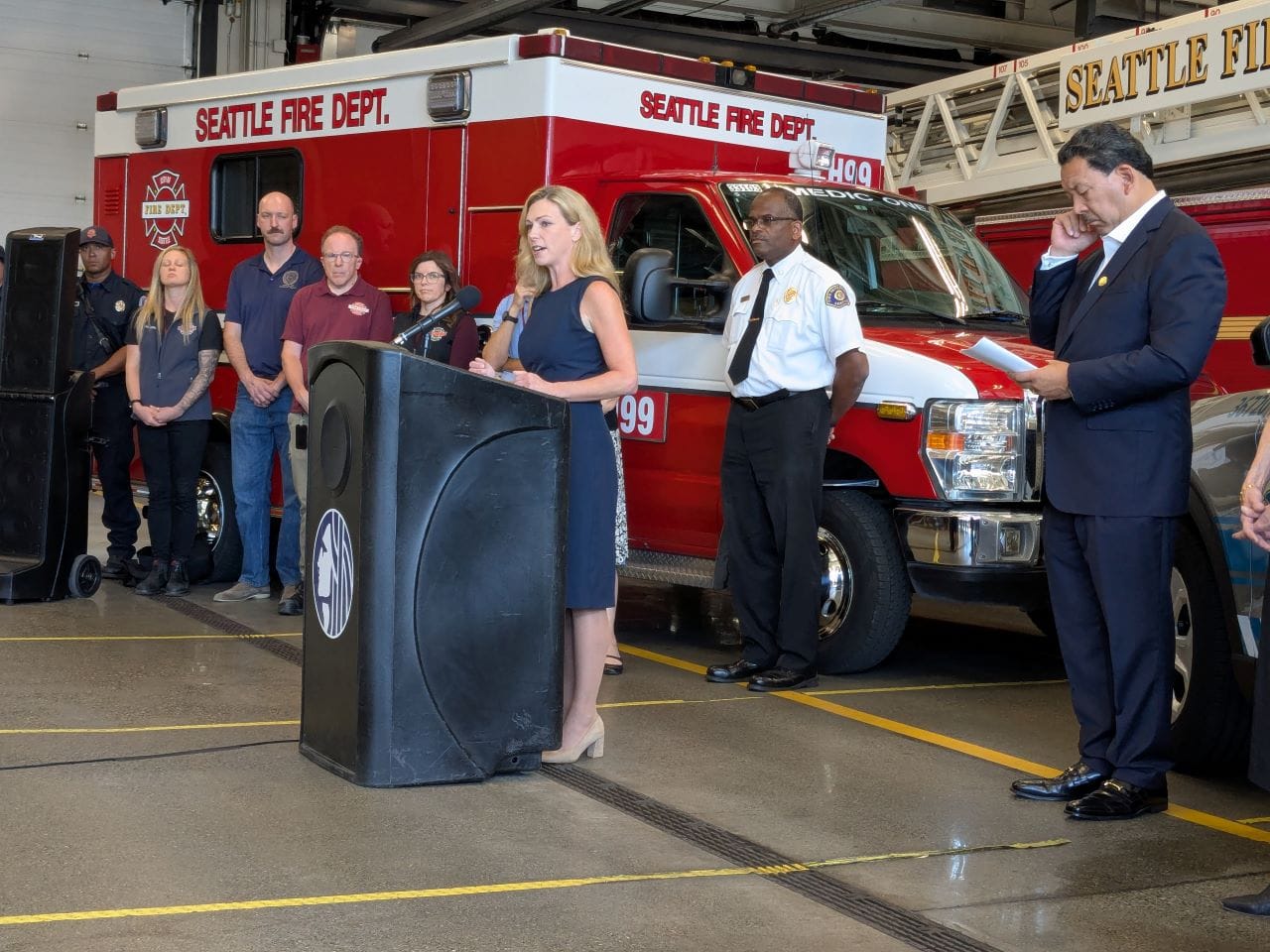 Barden stands at a lectern at a fire hall with a fire truck, fire chief, mayor, and CARE staff in the background.