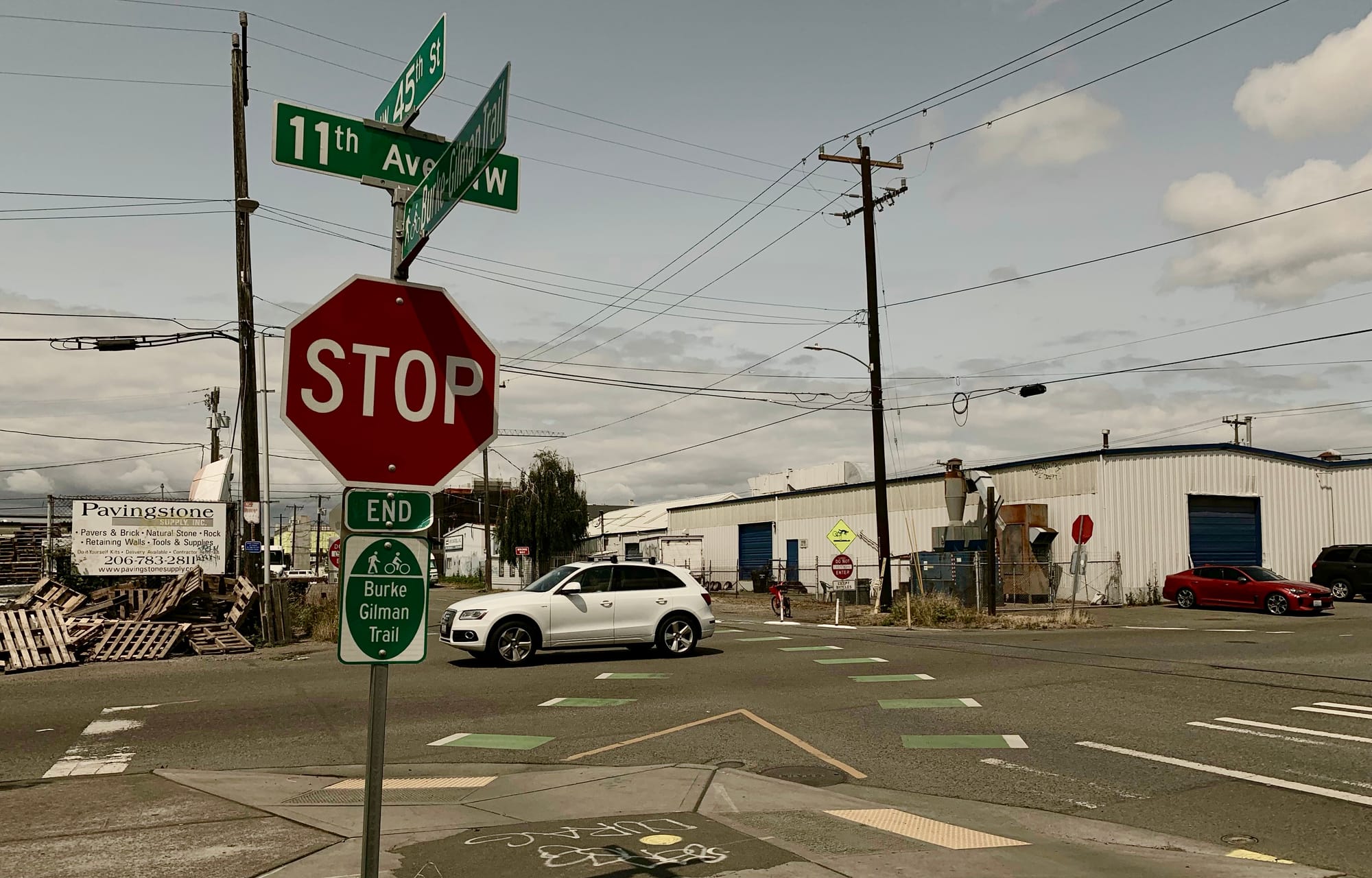 A photo showing a stop sign and a sign indicating the end of the Burke Gilman Trail. The street behind it shows a car turning into the intersection. 
