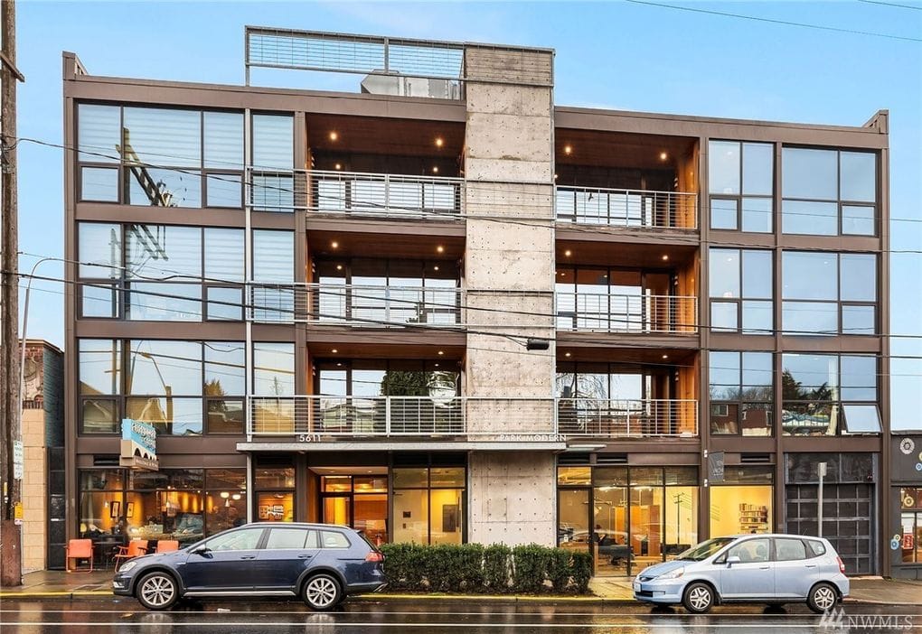 A photograph shows a modern mid-rise building with large glass windows as viewed from the street.