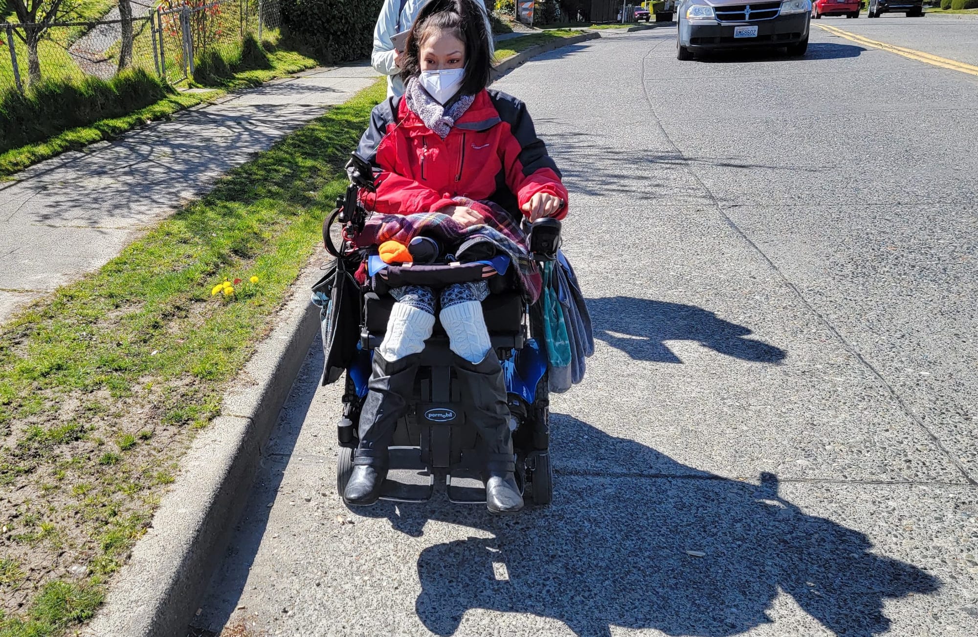 
                     A woman wearing a red jacket and face mask rides her power wheelchair in the road with a person walking behind her and a car
                     