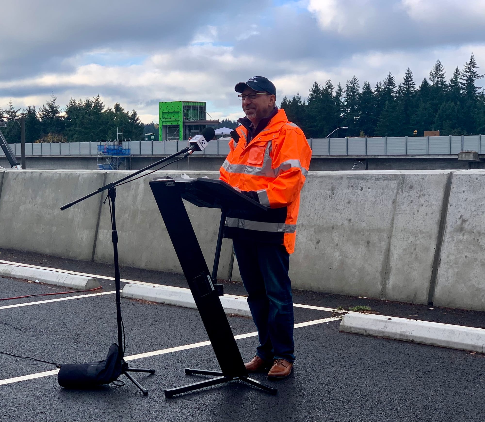 A photo of a man wearing an orange construction jacket and baseball hat standing at a podium. 