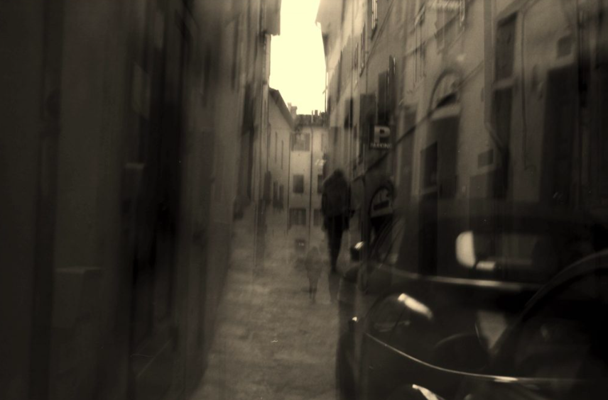 
                     A blurred black and white photo of a figure walking down a street and a car.
                     