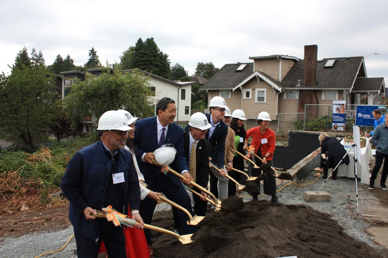 Wearing white hard hats, a line of officials pick up of a scoop of ceremonial dirt from a big pile