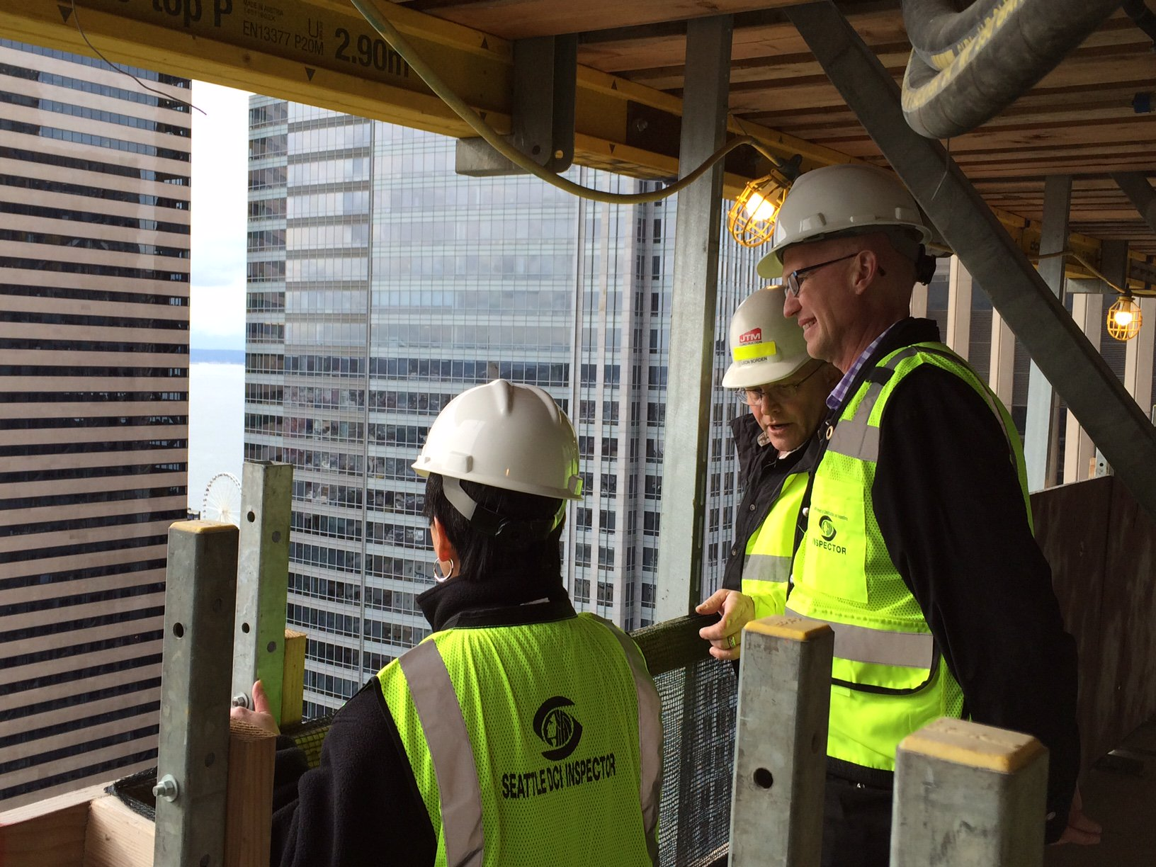 Torgelson and two others were yellow vests and white construction hats as they tour a downtown tower that is under construction. They stand at a window that looks out on other towers.