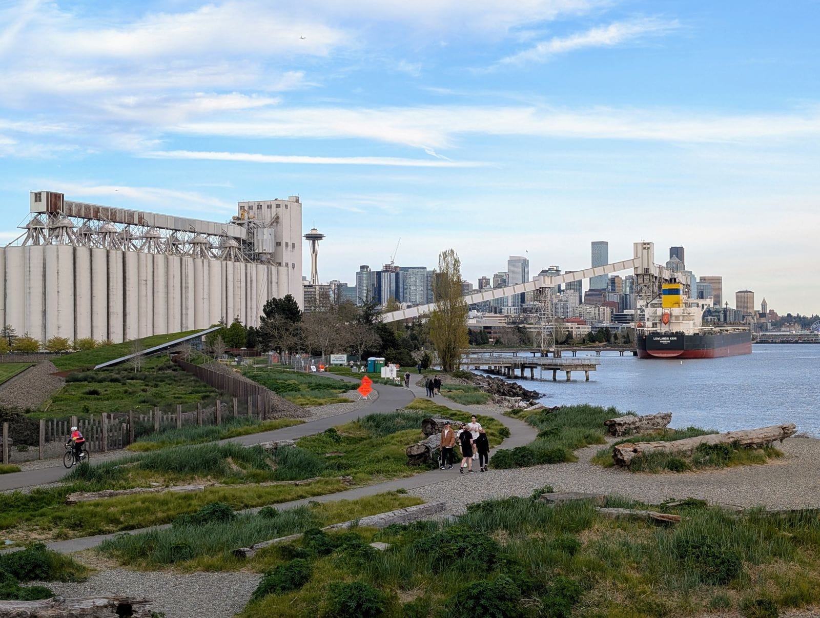 A grain elevator, the space needle, and skyline are visible in the distance. A smattering of joggers and walkers use the path.