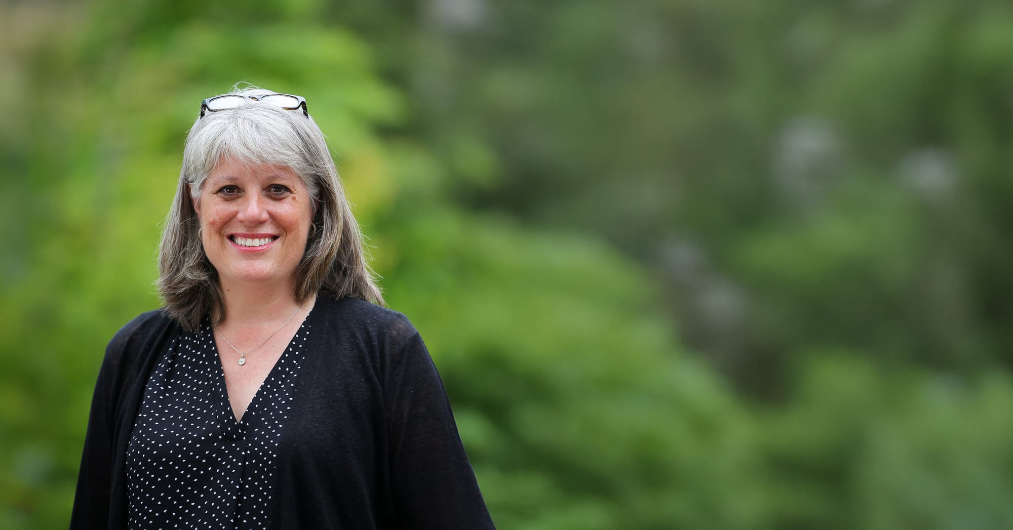 Balducci has her glasses up on her head and smiles. She stands in front of a forest.