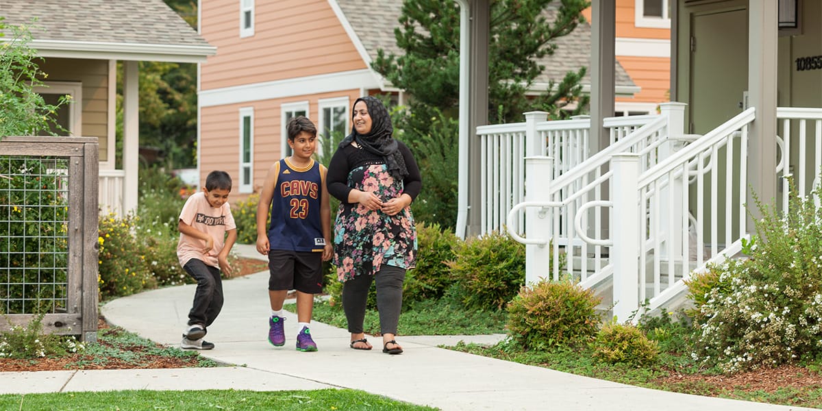A mother and her two sons walk on a pedestrian path between a row of townhomes. She wears a headscarf and her older son wears a Lebron James jersey. The younger is on a skateboard.