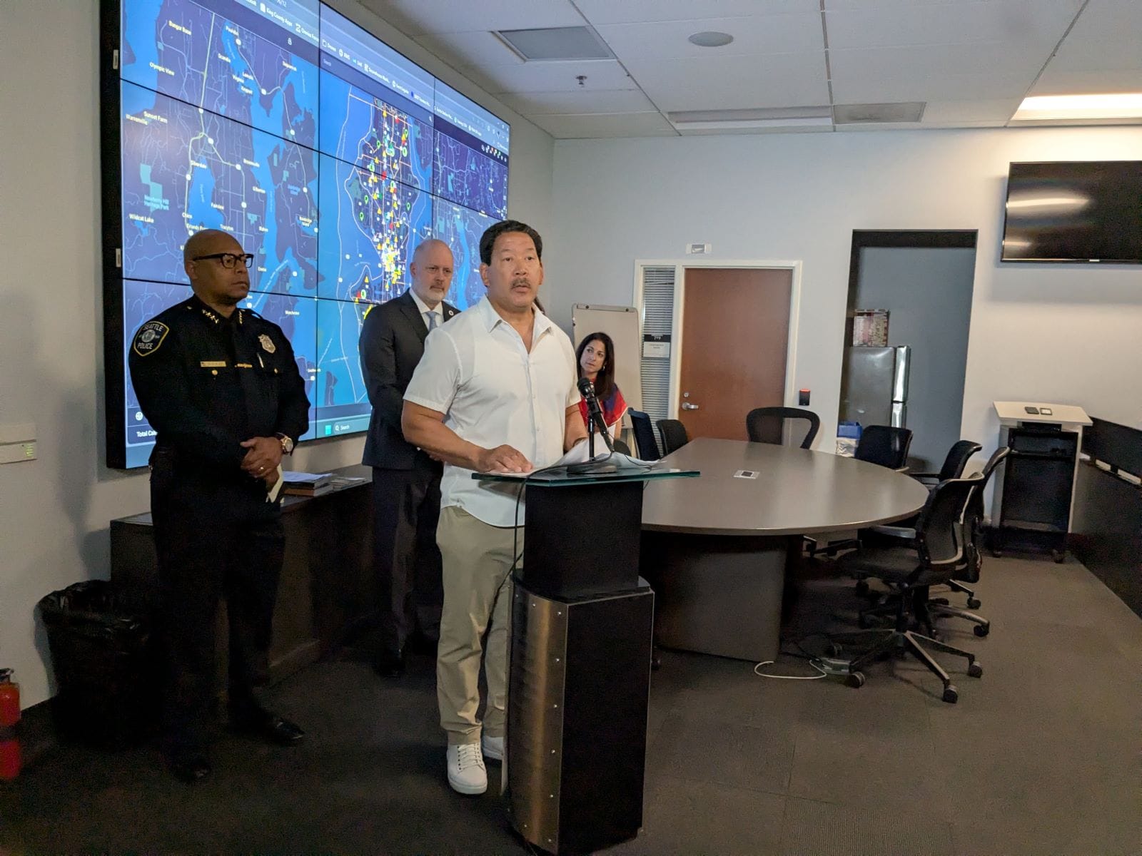 Mayor Bruce Harrell stands at a lectern flanked by Police Chief Shon Barnes and Councilmember Bob Kettle, who chairs the Public Safety Committee. A projection screen displays surveillance footage.