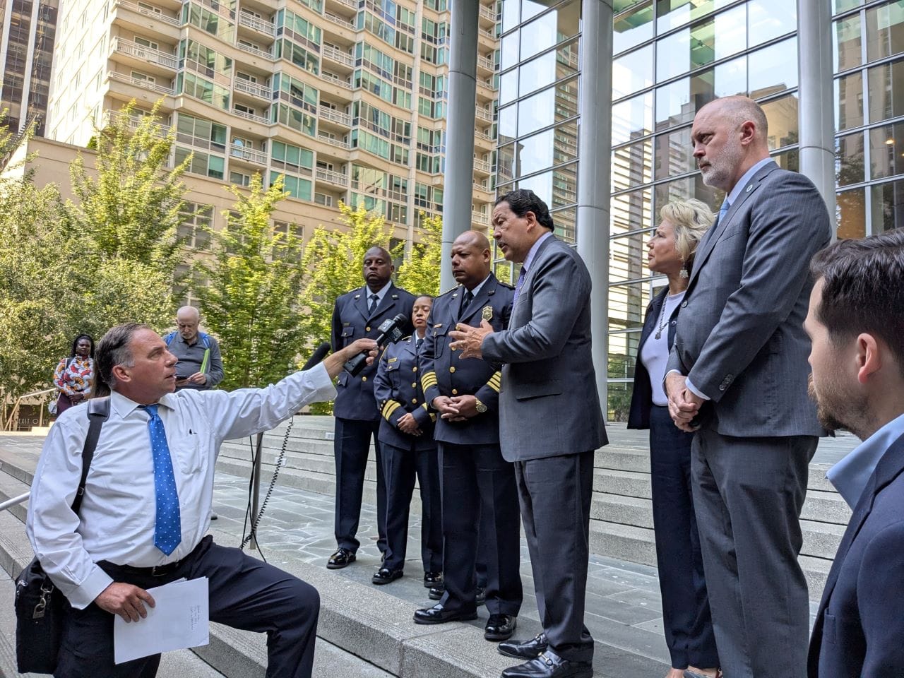 Officials including Bob Kettle and Debora Juarez and SPD top brass stand on the steps of Seattle police headquarters with a reporter outstretching an arm holding a mic to interview them