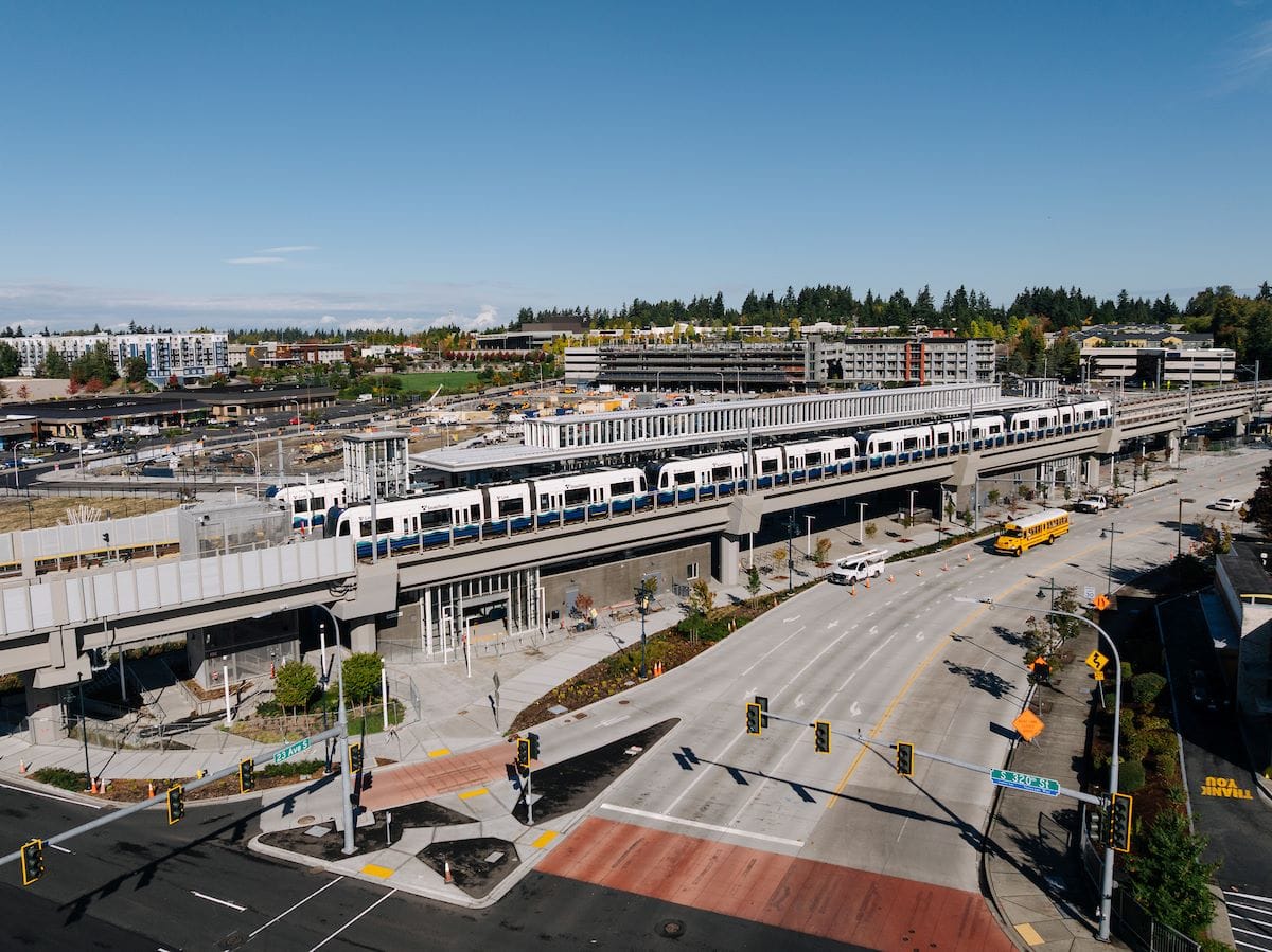 A drone photo shows a four-car train at Federal Way Station during testing. Some mid-rise buildings are off in the distance.