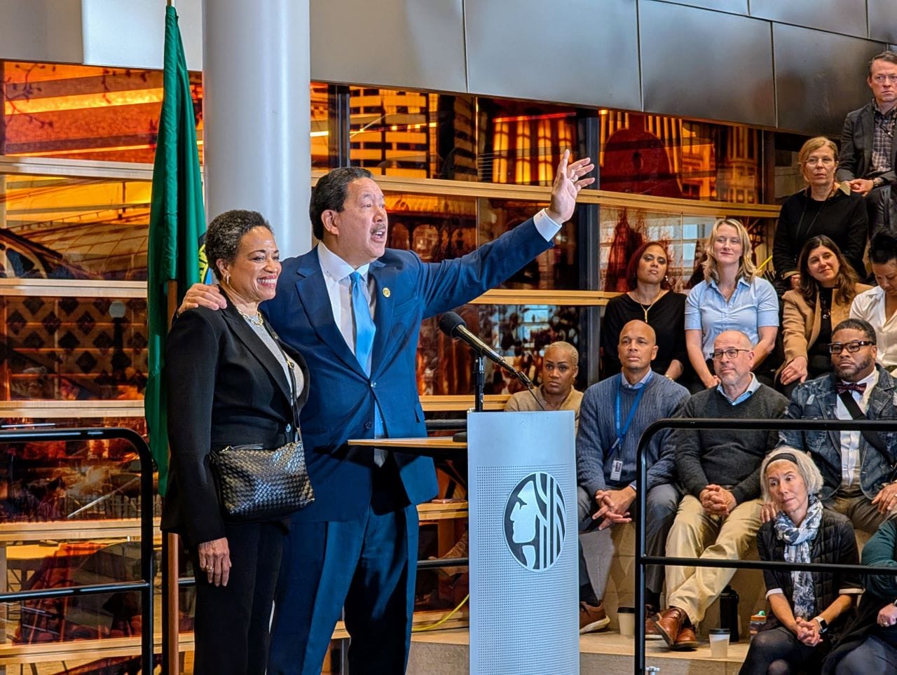 
                     Harrell waves goodbye at the end of his speech with his wife on his arm. They stand at a lectern in the lobby of city hall.
                     