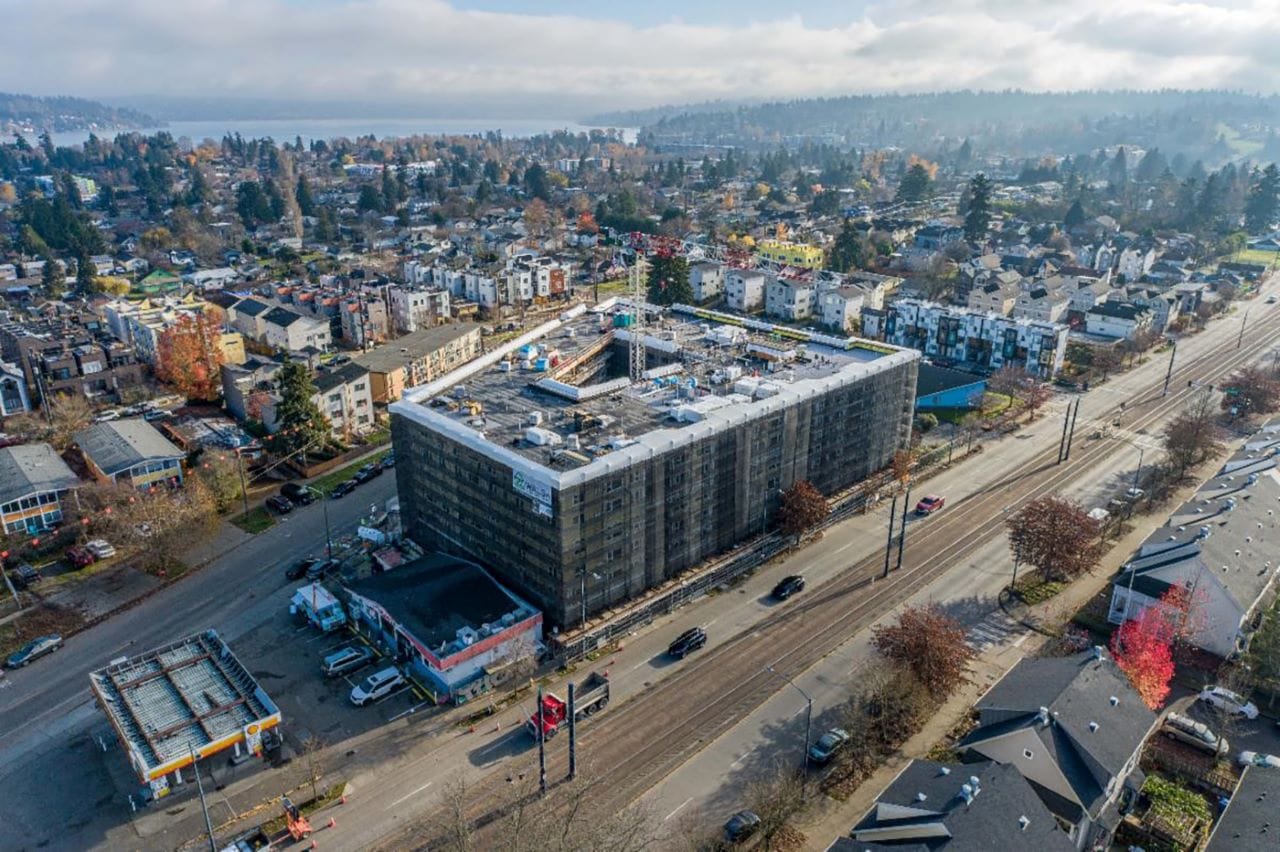 A yellow construction wrap is around the six-story building. Townhomes, treetops, and Lake Washington are in the background.