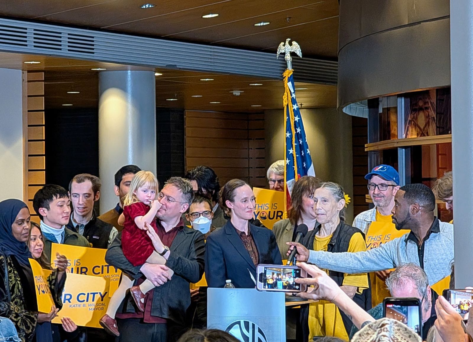 
                     Wilson supporters hold yellow "This is your city" signs. She stands at the lectern and and a man adjust the mic to her height
                     