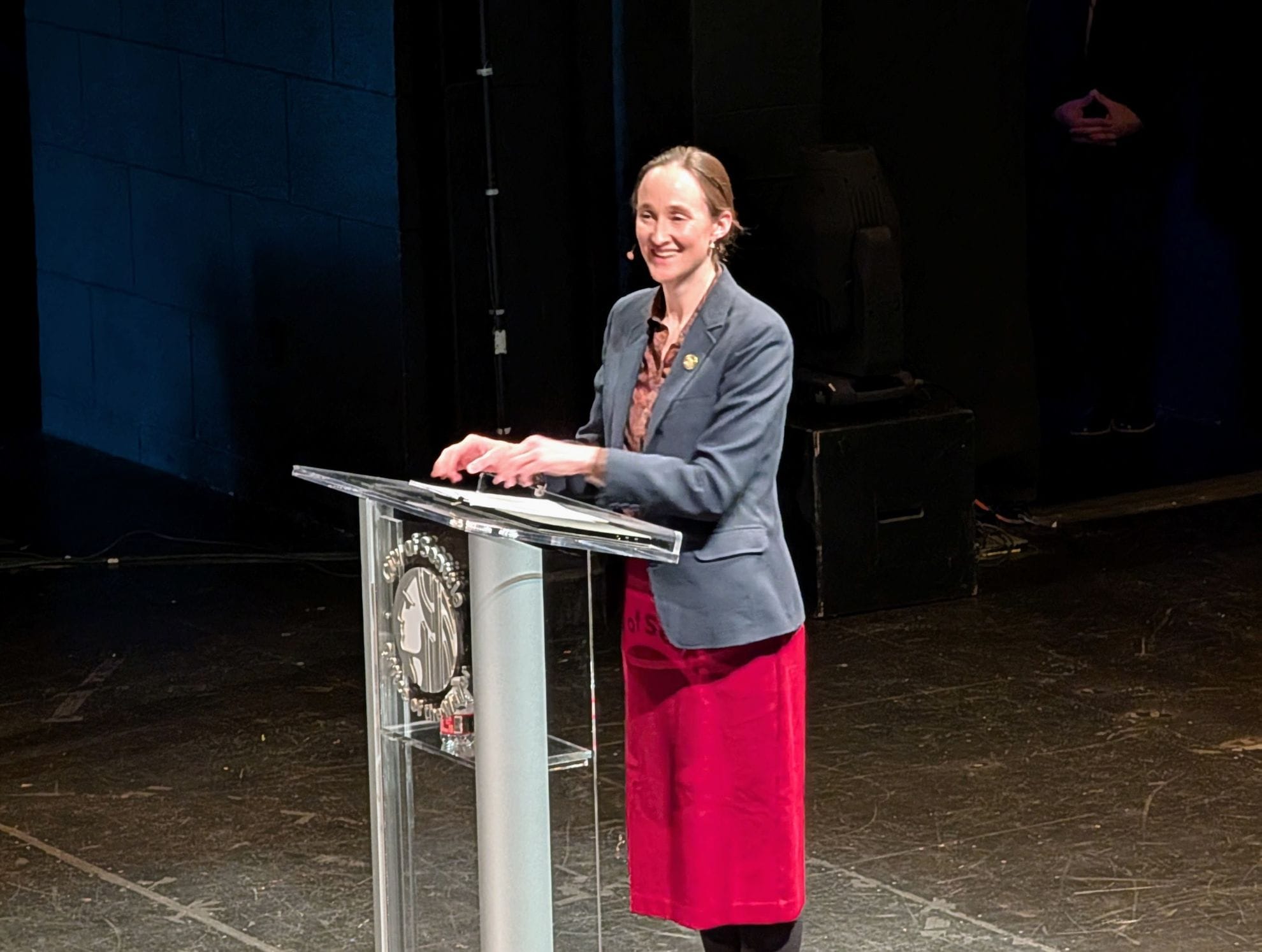 
                     Wilson smiles and stands at a lectern with the City seal. She wears a red skirt and gray blazer.
                     