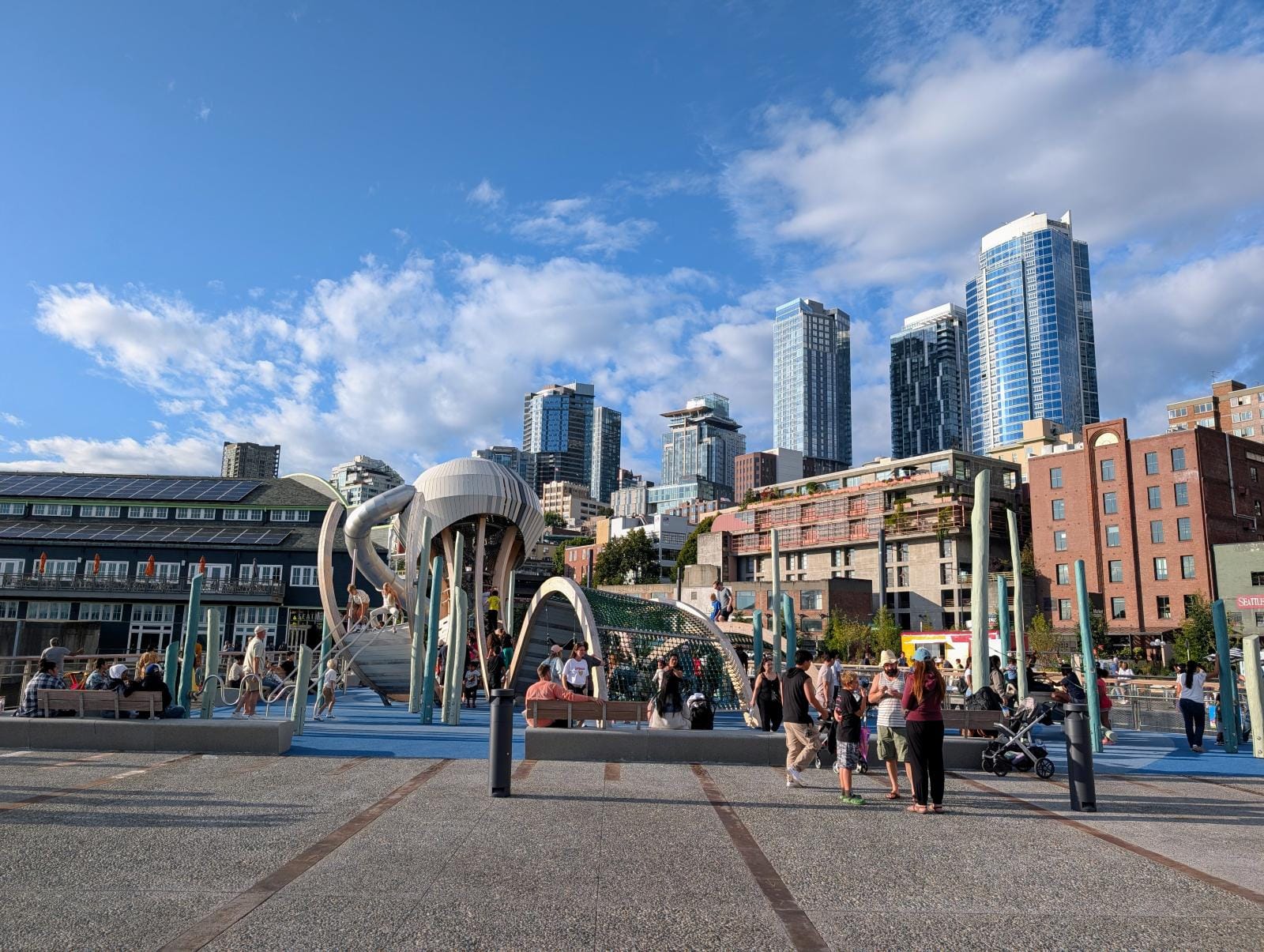 The Seattle skyline stands in the background at a crowded playground on a sunny summer day.