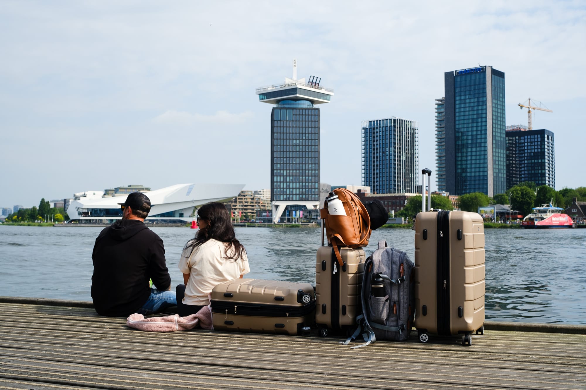 Couple sitting on a wooden dock by the IJ river in Amsterdam with suitcases and bags, looking towards the Noord skyline